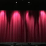 Closed Red theater curtains on stage with lights from above partially lighting the curtains